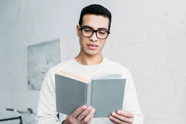 Joven mixto raza serio hombre en blanco sudadera y gafas lectura libro - foto de stock