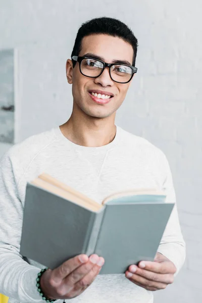 Sonriente mestizo joven en gafas sosteniendo libro y mirando a la cámara - foto de stock