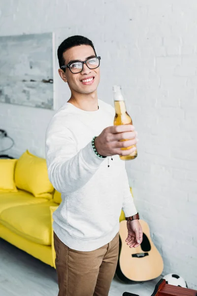 Sonriente guapo mixto raza hombre celebración botella de cerveza y mirando cámara - foto de stock