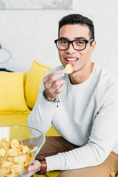 Красивий молодий змішаний гоночний чоловік сидить на жовтому дивані і їсть чіпси — стокове фото