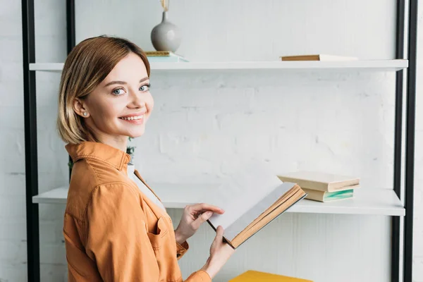 Smiling pretty girl in orange shirt holding book while standing by shelving rack — Stock Photo