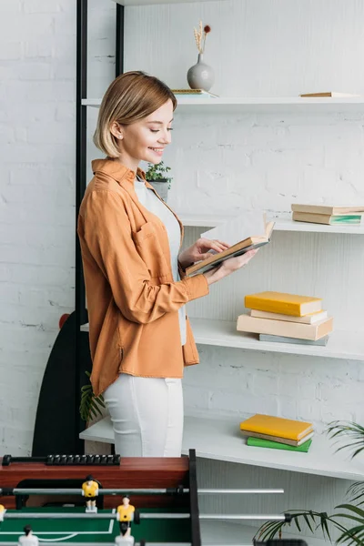 Beautiful girl reading book while standing by shelving rack — Stock Photo