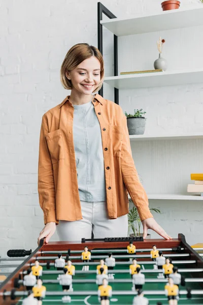 Smiling pretty girl in orange shirt and white jeans standing by football table — Stock Photo