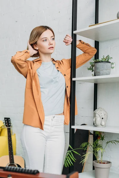 Chica atractiva en camisa naranja y pantalones vaqueros blancos de pie por estantería y el pelo conmovedor - foto de stock
