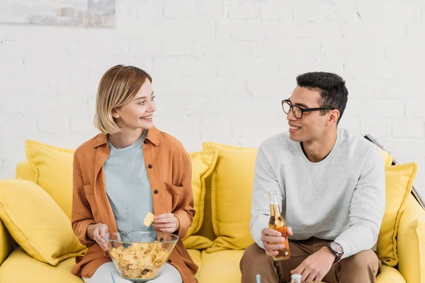 Interrassische Paare reden und genießen Snacks und Getränke, während sie auf dem gelben Sofa sitzen — Stockfoto