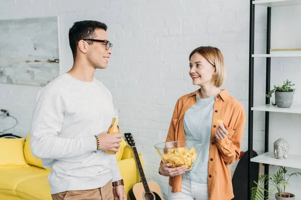 Interracial couple chatting and enjoying snacks and drinks while standing in living room — Stock Photo