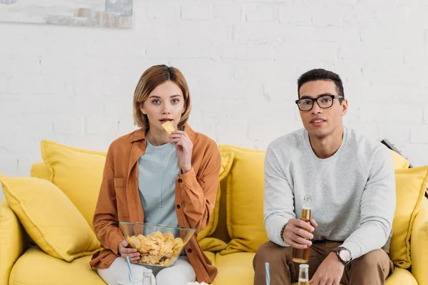 Pareja interracial disfrutando de aperitivos y bebidas mientras se sienta en un sofá amarillo - foto de stock