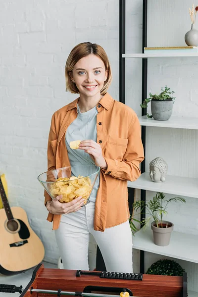 Sonriente chica bonita sosteniendo tazón de patatas fritas y mirando a la cámara - foto de stock