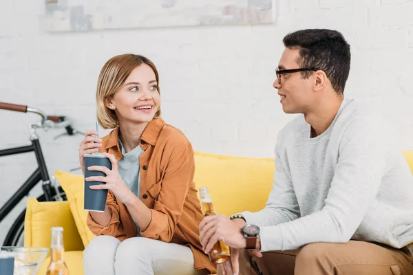 Feliz interracial pareja sentado en amarillo sofá y disfrutar de bebidas - foto de stock