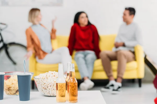 Selektiver Schwerpunkt Tisch mit Snacks und Getränken, multikulturelle Freunde sitzen auf gelbem Sofa — Stockfoto