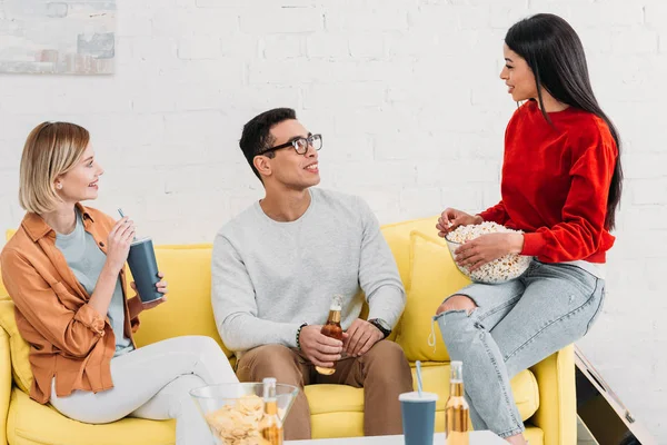 Amis multiculturels souriants dégustant des boissons et des collations tout en étant assis sur un canapé jaune — Photo de stock