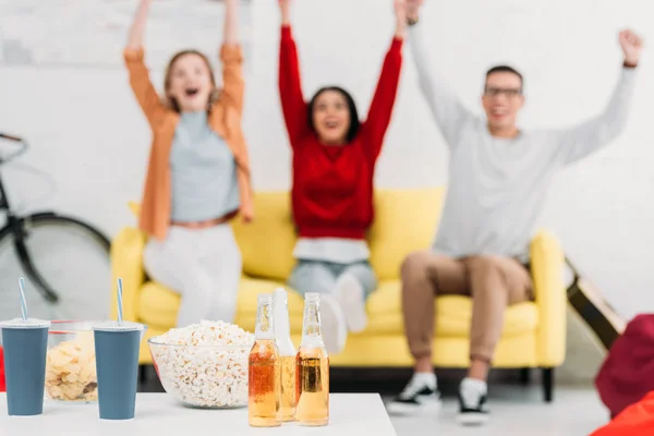 Enfoque selectivo de mesa con aperitivos y bebidas, amigos multiculturales sentados en un sofá amarillo y levantando las manos - foto de stock