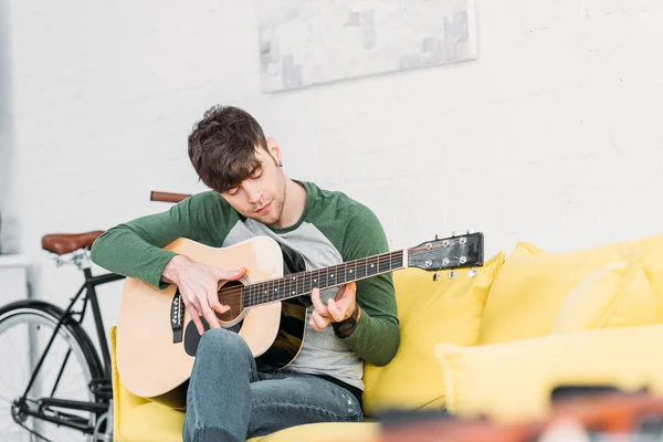 Schöner junger Mann spielt Akustikgitarre, während er auf gelbem Sofa sitzt — Stockfoto