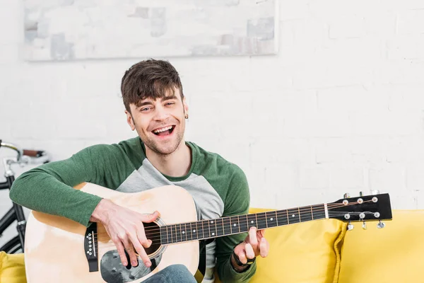 Молодий чоловік грає на акустичній гітарі, сидячи на жовтому дивані — стокове фото