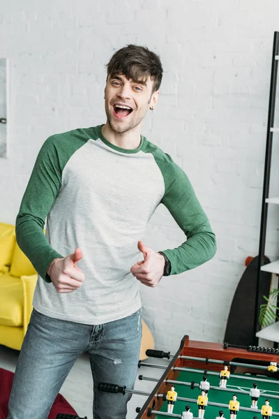 Handsome young man standing by football table and showing thumbs up — Stock Photo