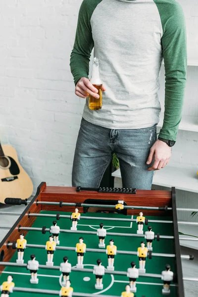 Vista parcial del hombre de pie junto a la mesa de fútbol y sosteniendo la botella de cerveza - foto de stock
