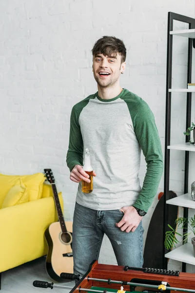 Souriant jeune homme debout dans le salon et tenant une bouteille de bière — Photo de stock