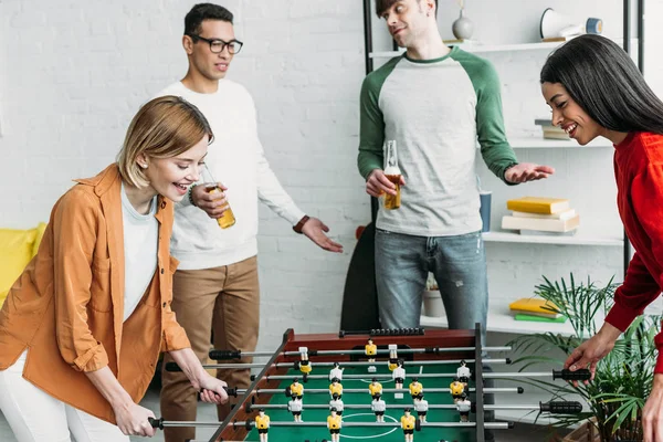 Sonrientes chicas multiculturales jugando al futbolín mientras los hombres hablan y beben cerveza - foto de stock