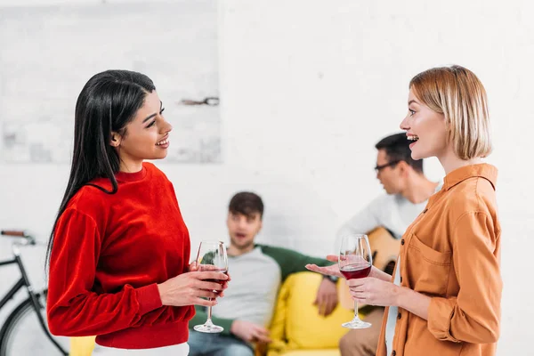 Amigos multiculturales sosteniendo gafas y hablando mientras los hombres se sientan en el sofá amarillo - foto de stock