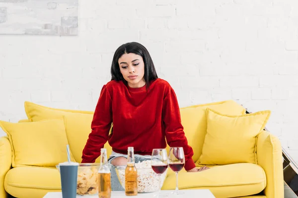 Довольно африканская американская девушка сидит на желтом диване рядом со столом с напитками и закусками — стоковое фото