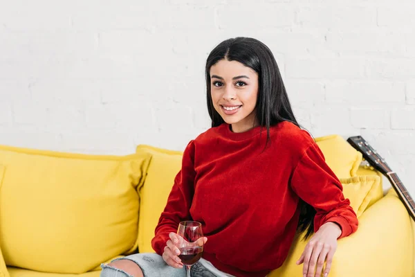 Chica afroamericana sonriente sentada en un sofá amarillo y sosteniendo un vaso de vino - foto de stock