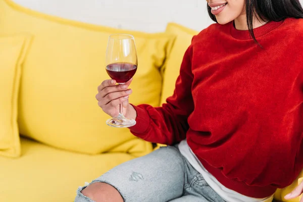 African american woman sitting on yellow sofa and holding glass of wine — Stock Photo