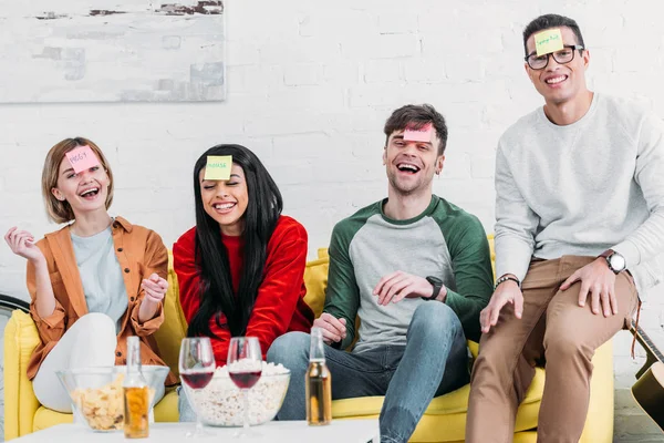 Laughing multicultural friends with funny nickname labels on foreheads sitting on yellow sofa — Stock Photo