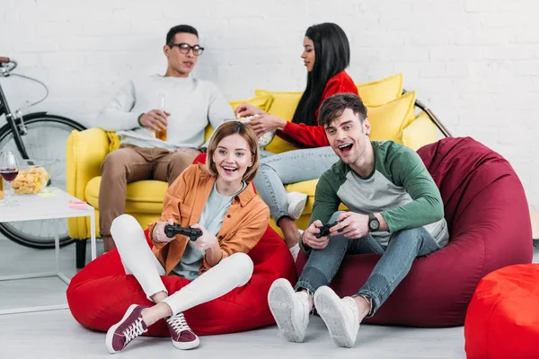 Amigos multiculturales felices jugando videojuegos y disfrutando de bebidas y aperitivos en la fiesta en casa - foto de stock