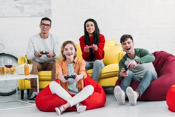 Amigos multiculturales emocionados jugando videojuegos y disfrutando de aperitivos y bebidas en la fiesta en casa - foto de stock