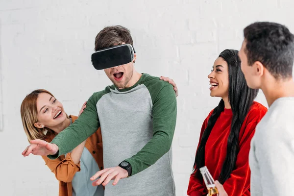 Amigos multiétnicos felices riéndose del hombre usando auriculares de realidad virtual - foto de stock