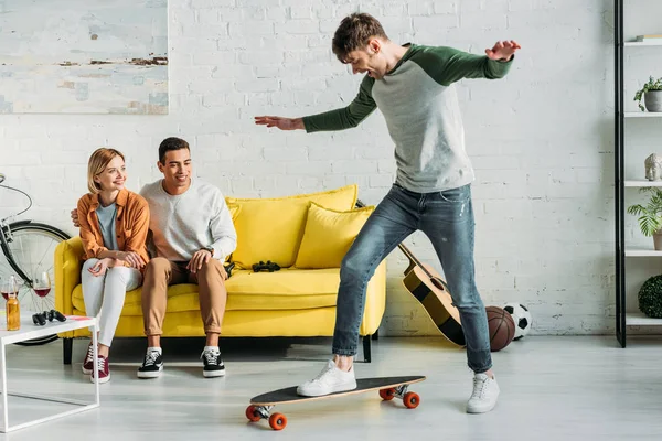 Multicultural friends sitting on yellow sofa and looking at friend riding longboard — Stock Photo
