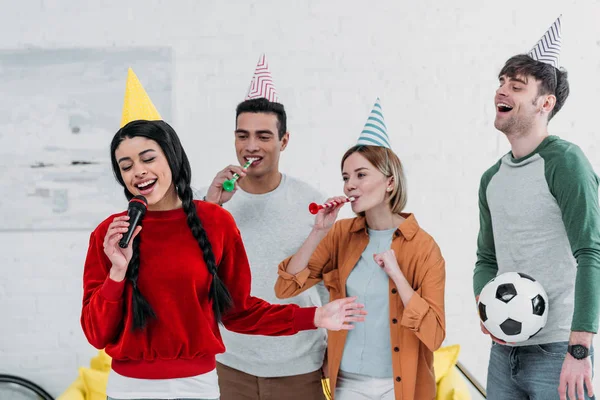 Amigos multiculturales en coloridos sombreros de papel cantando karaoke en casa fiesta - foto de stock