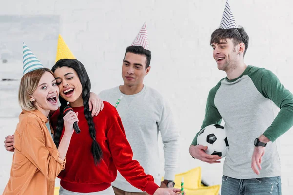 Amis multiculturels en chapeaux de papier coloré chantant karaoké à la fête à la maison — Photo de stock