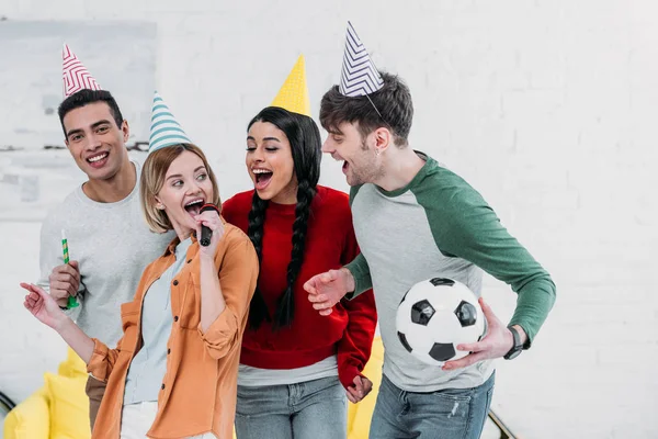 Amis multiethniques joyeux dans des chapeaux en papier multicolore s'amuser à la fête à la maison et chanter karaoké — Photo de stock