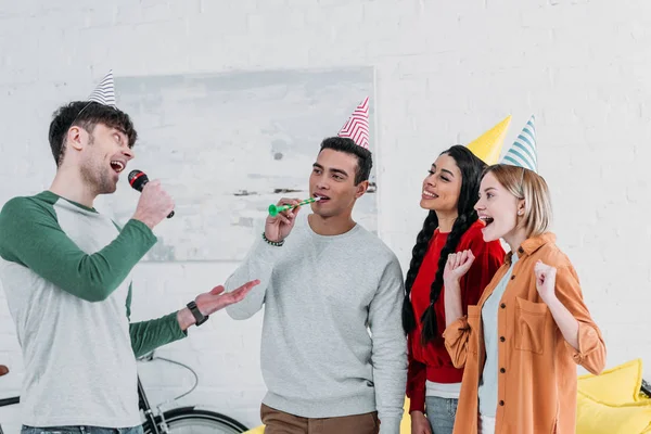 Amigos multiculturales felices en coloridos sombreros de papel cantando karaoke en la fiesta en casa - foto de stock