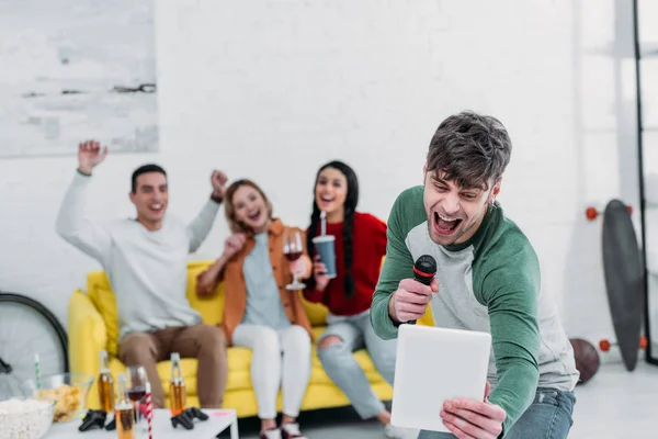 Jóvenes alegres cantando karaoke mientras amigos multiétnicos disfrutan de bebidas y aperitivos - foto de stock