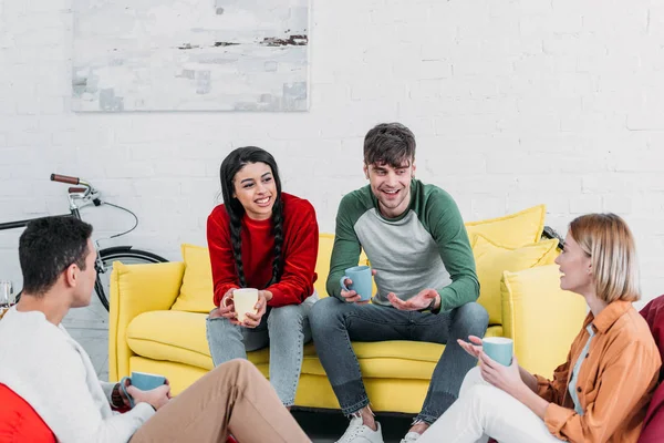 Sonrientes amigos multiculturales tomando café y hablando mientras están sentados en la sala de estar - foto de stock