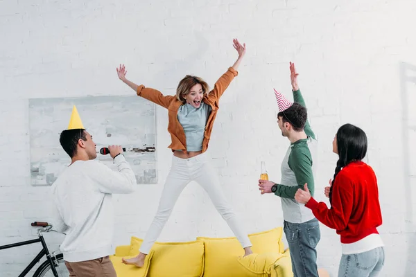 Cheerfull girl jumping on yellow sofa while multicultural friends singing karaoke and dancing — Stock Photo