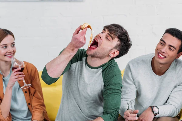 Jovem alegre comendo pizza enquanto amigos multiculturais gostando de bebidas — Fotografia de Stock