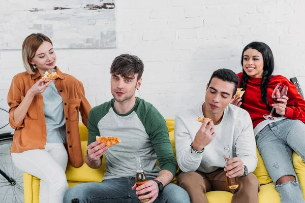 Fröhliche multikulturelle Freunde, die Pizza essen und Getränke genießen, während sie zusammen auf dem gelben Sofa sitzen — Stockfoto