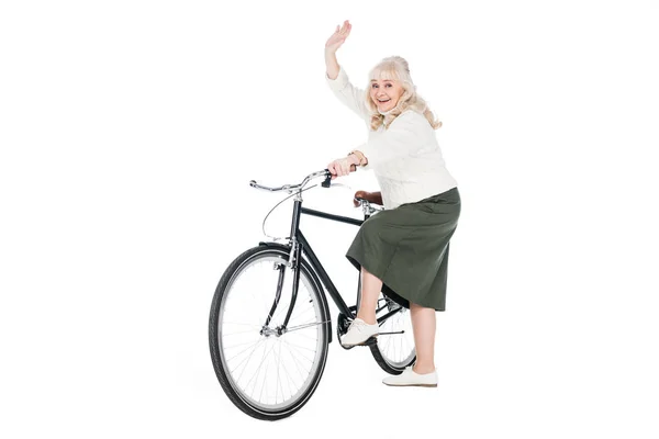 Весела старша жінка їде на велосипеді і махає рукою ізольовано на білому — стокове фото
