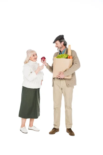 Allegro uomo anziano mostrando mela alla moglie, mentre tenendo sacchetto di carta con generi alimentari isolati su bianco — Foto stock