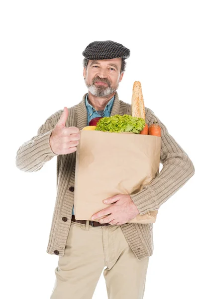 Allegro uomo anziano tenendo sacchetto di carta con generi alimentari e mostrando pollice su isolato su bianco — Foto stock