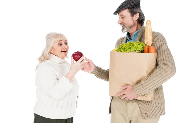 Senior man showing apple to cheerful wife while holding paper bag with groceries isolated on white — Stock Photo