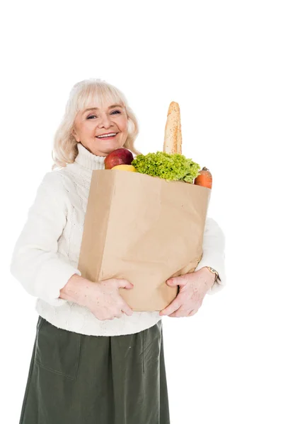 Alegre mujer jubilada sosteniendo bolsa de papel con comestibles aislados en blanco - foto de stock
