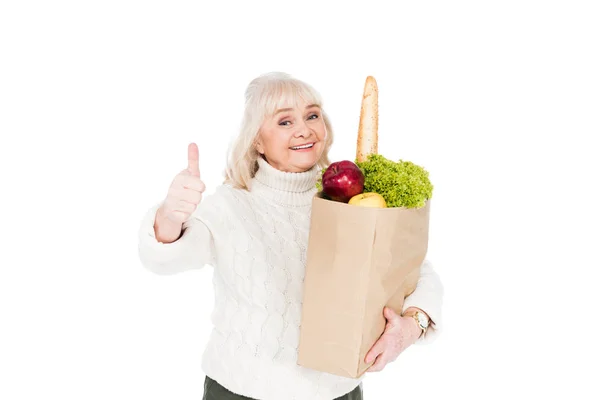 Cheerful retired woman holding paper bag with groceries and showing thumb up sign isolated on white — Stock Photo