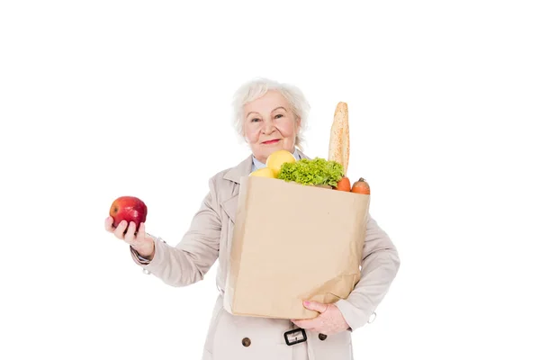 Весела жінка з сірим волоссям тримає яблуко і паперовий мішок з продуктами харчування ізольовані на білому — стокове фото