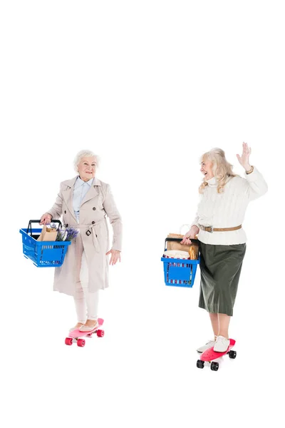 Happy senior woman riding penny board and waving hand to friend holding shopping basket idolated on white — Stock Photo