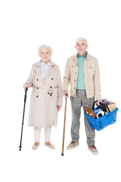 Alegre pareja jubilada sosteniendo cestas de compras y bastones mientras está de pie aislado en blanco - foto de stock