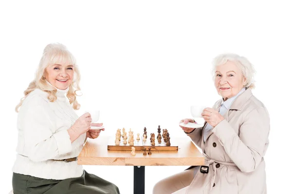 Femmes à la retraite tenant des tasses avec des boissons tout en jouant aux échecs isolés sur blanc — Photo de stock